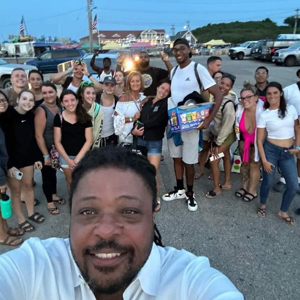 Large group poses for a cheerful waterfront parking-lot selfie, with boats, flags, cars, and buildings in the background.