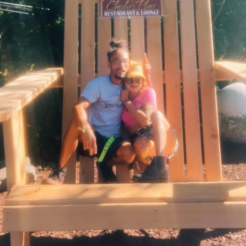 A couple sits on an oversized wooden chair beneath a Charlie’s Place Restaurant & Lounge sign, surrounded by leafy trees.