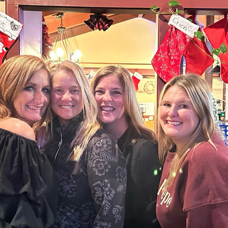 Four smiling women pose together inside a warmly decorated restaurant with Christmas stockings, plants, and a colorful game board.