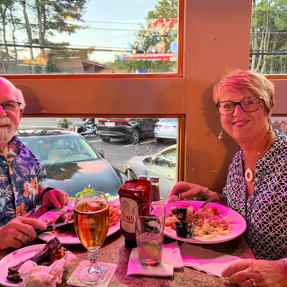 Older couple smiling at a restaurant table with plates of food, drinks, and a street view through the window.