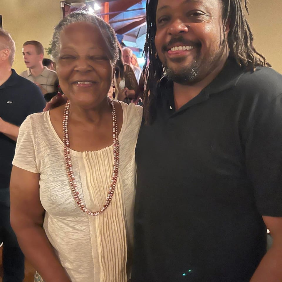 Two smiling people pose together at a lively indoor restaurant gathering, surrounded by other guests.