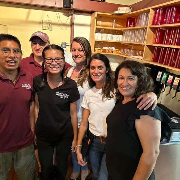 Six smiling restaurant staff members pose together behind the counter, surrounded by drink dispensers, cups, and colorful supplies.