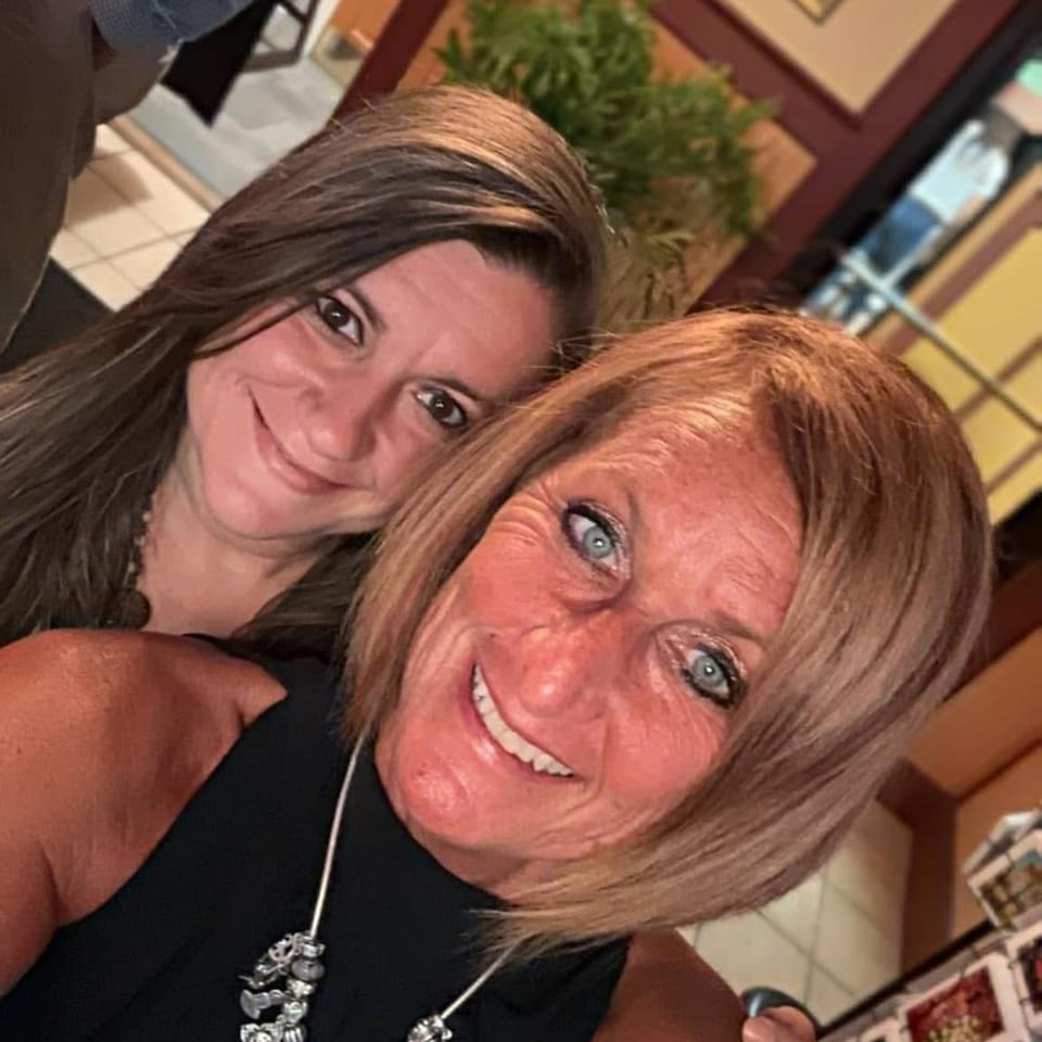 Two smiling women pose together indoors, with framed artwork, a potted plant, and restaurant furnishings in the background.