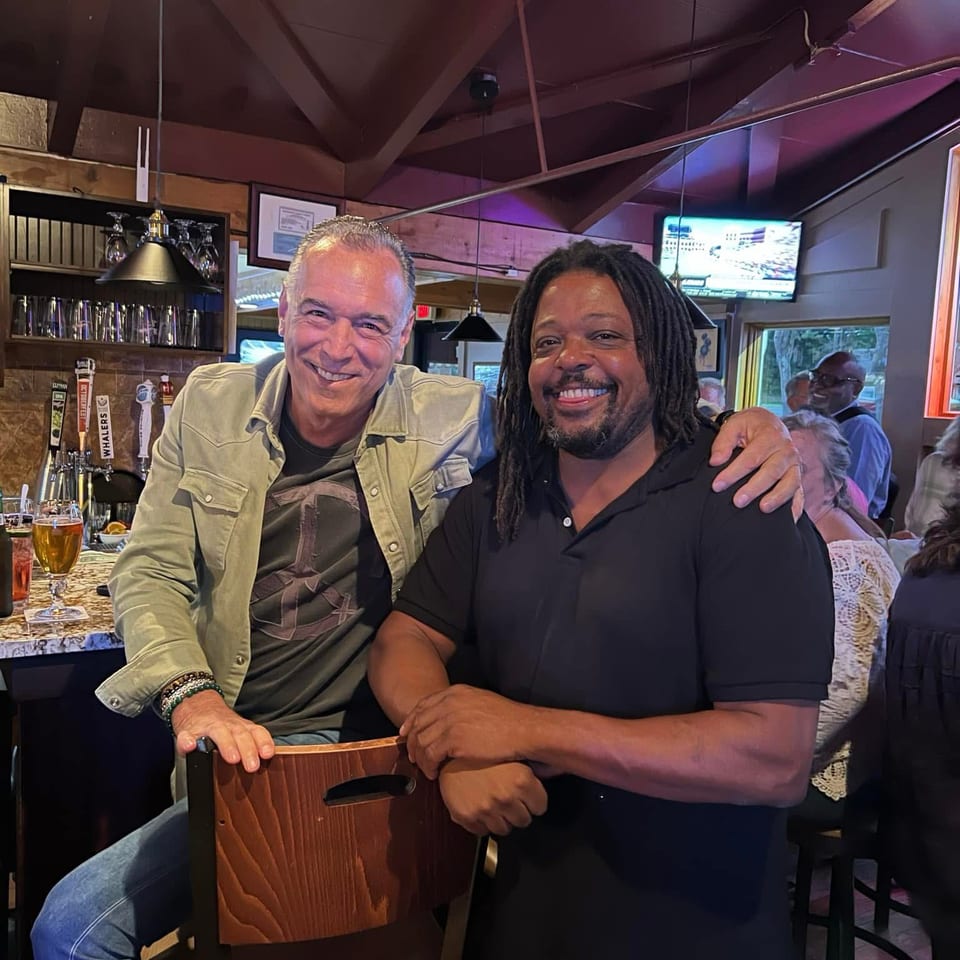 Two smiling men pose together inside a warmly lit restaurant bar, with beer taps, glasses, diners, and a television in the background.