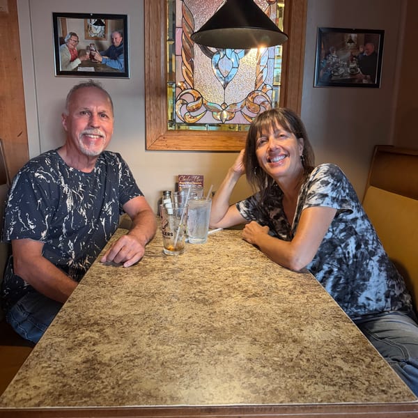Two diners smile at a restaurant table with drinks, beneath a pendant lamp and decorative stained-glass window.