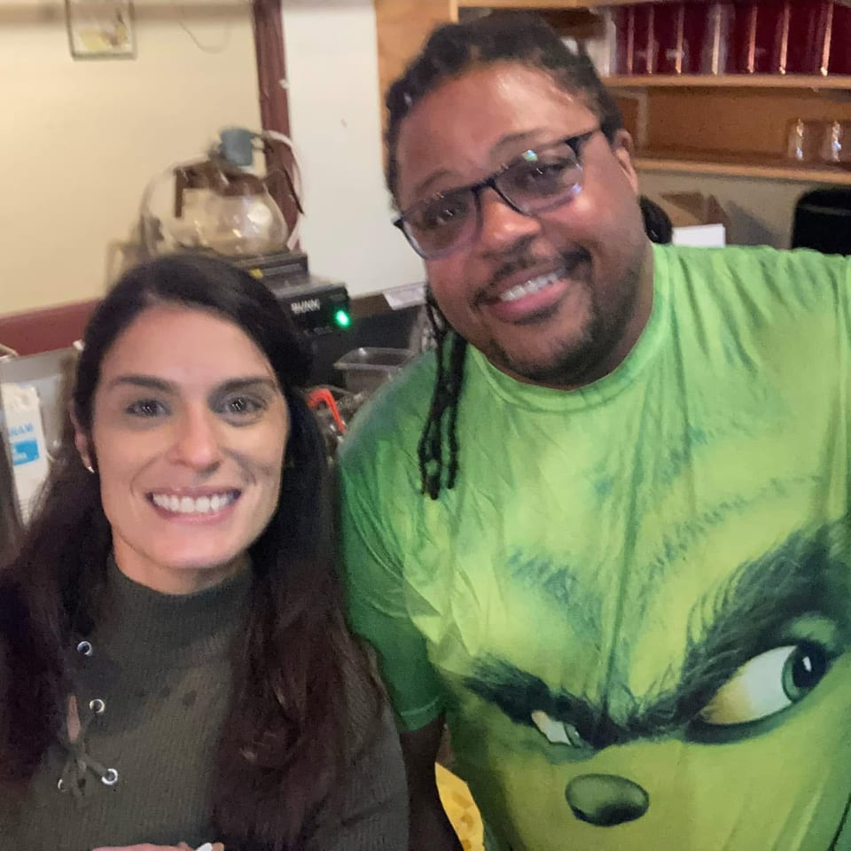 Smiling pair pose for a selfie inside a restaurant, with shelves, beverage dispensers, and kitchen equipment behind them.