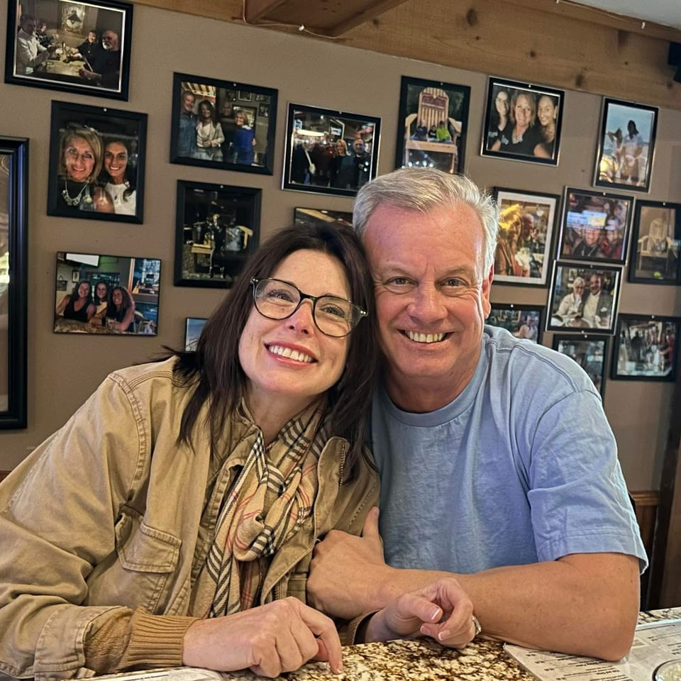 Smiling couple poses together at a restaurant counter, with framed photographs and memorabilia covering the wall behind them.