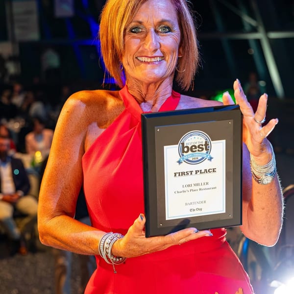 Smiling bartender Lori Miller in a red dress holds a first-place award for Charlie’s Place Restaurant at a lively event.