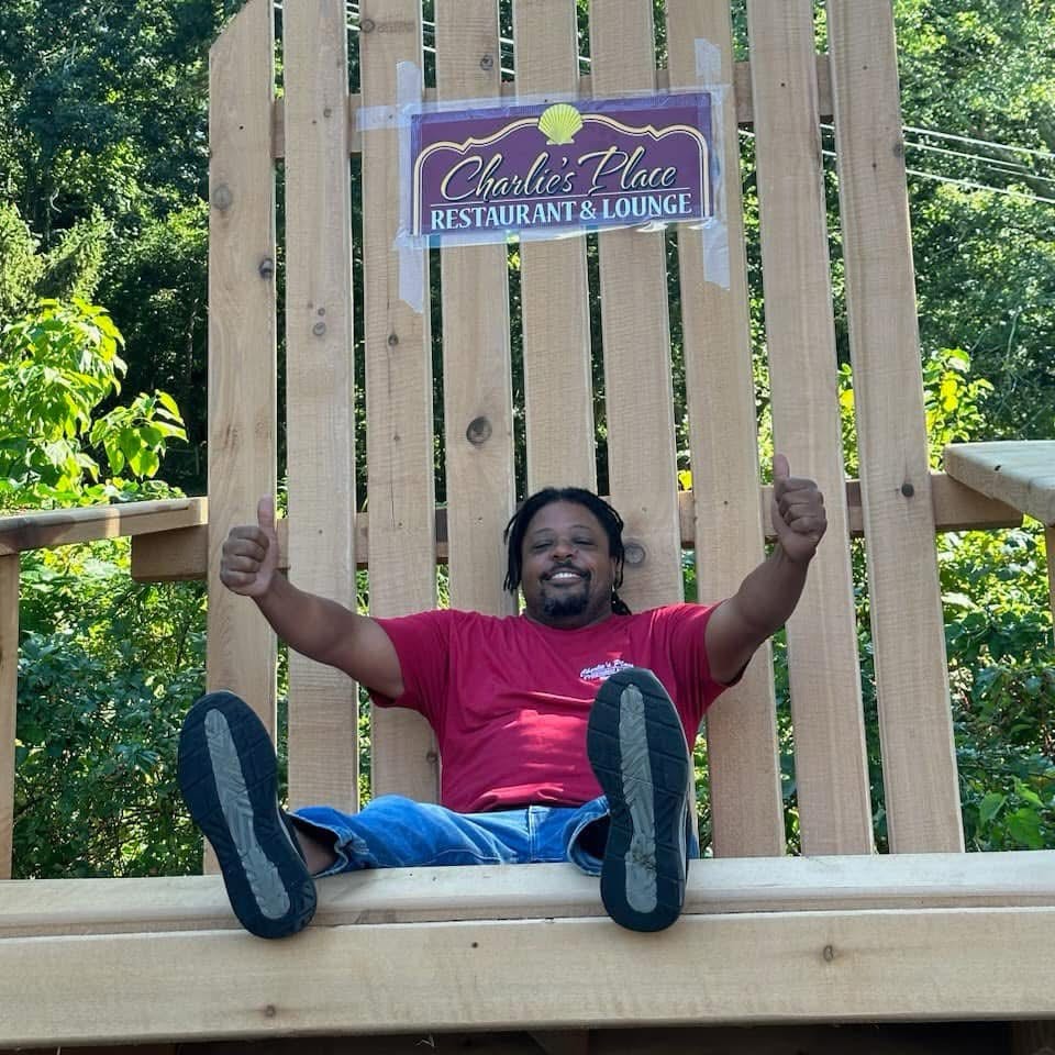 Man giving two thumbs up beneath a Charlie’s Place Restaurant & Lounge sign on a wooden outdoor deck surrounded by trees.