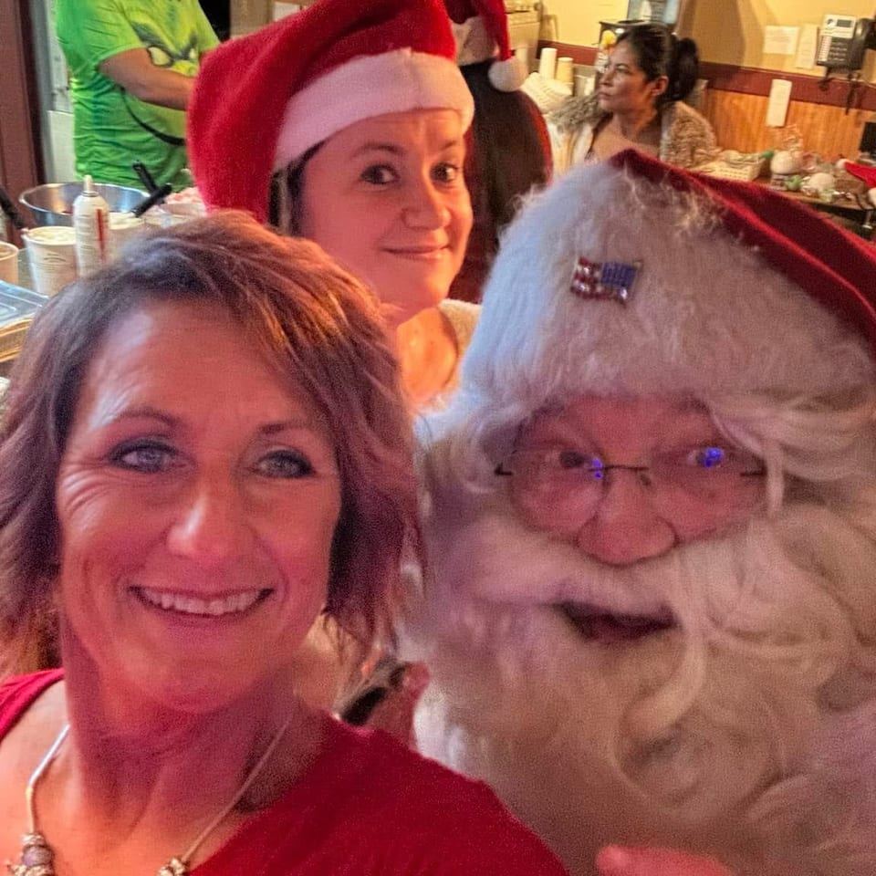 Three smiling women pose for a festive selfie inside a restaurant, with one wearing a Santa hat and buffet dishes visible behind them.