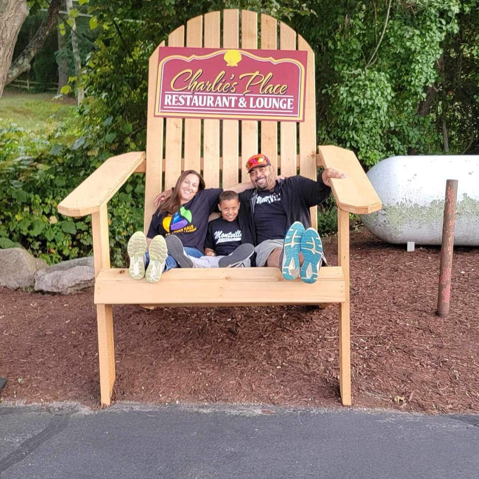 Three people smile while sitting in a giant wooden chair beneath a Charlie’s Place Restaurant & Lounge sign.