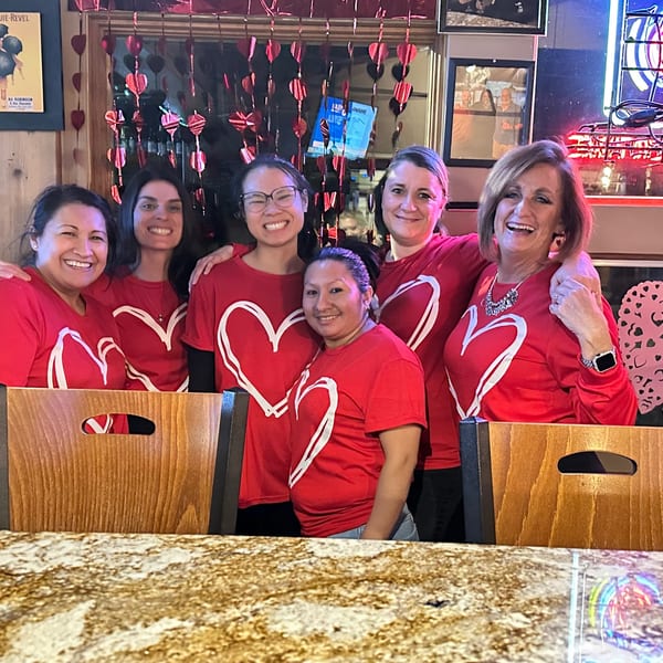 Five smiling women in matching red heart shirts pose together inside a warmly decorated restaurant.