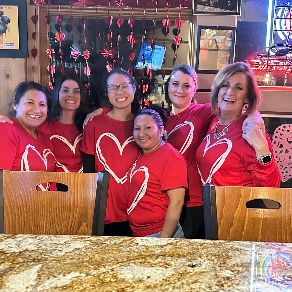 Five smiling women in matching red heart shirts pose together inside a warmly decorated restaurant.