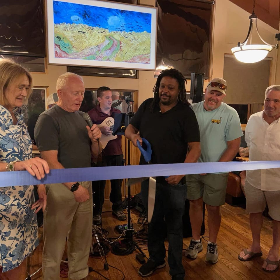 Group gathers indoors for a ribbon-cutting ceremony, with a musician, microphone, blue ribbon, and colorful landscape painting.