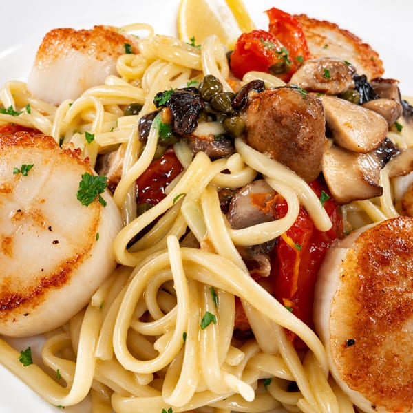 Seared scallops and pasta with mushrooms, tomatoes, and capers, garnished with parsley and lemon.