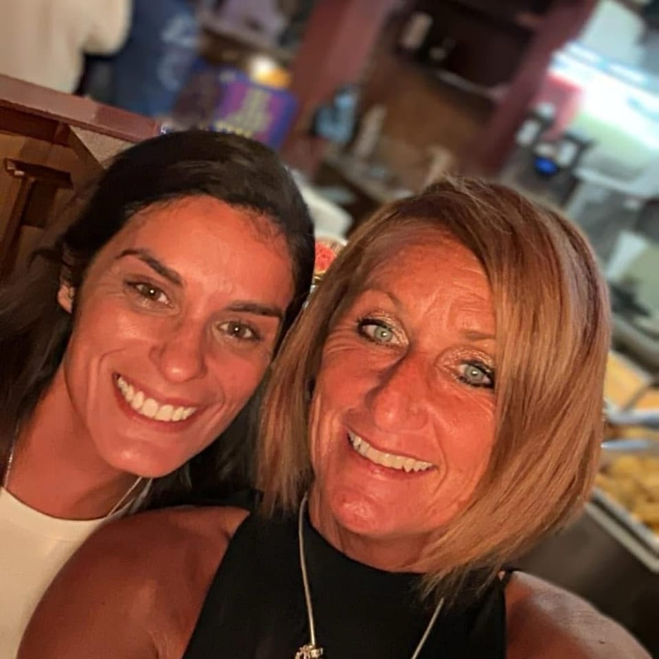 Two smiling women pose for a selfie inside a lively restaurant, with tables, diners, and colorful screens visible behind them.