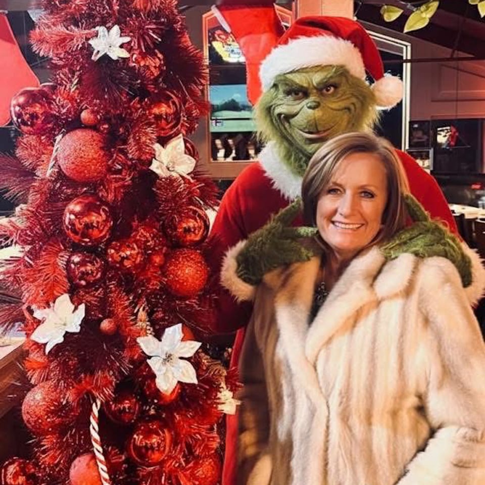 Woman in a fur coat poses beside a Grinch in a Santa hat and a decorated red Christmas tree indoors.