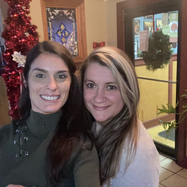 Two smiling women pose together inside a warmly decorated restaurant near a red Christmas tree and festive window.