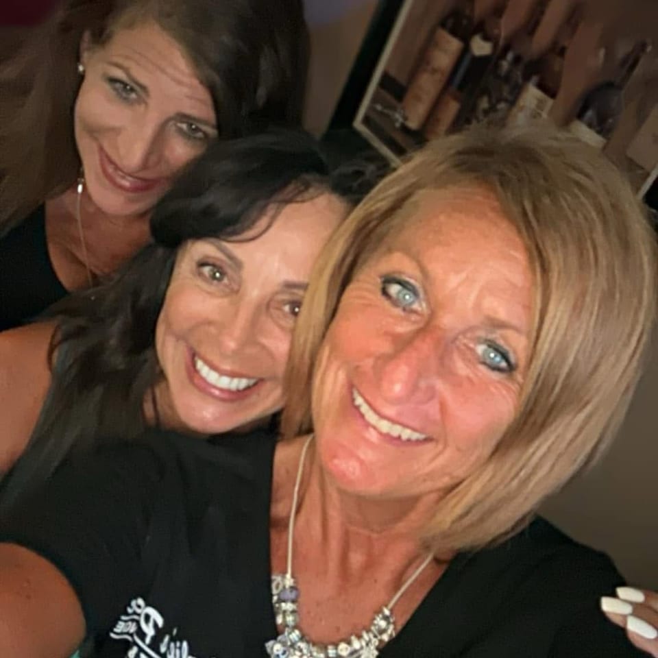 Three smiling women pose for a selfie in a dimly lit restaurant, with framed artwork and a wine display behind them.