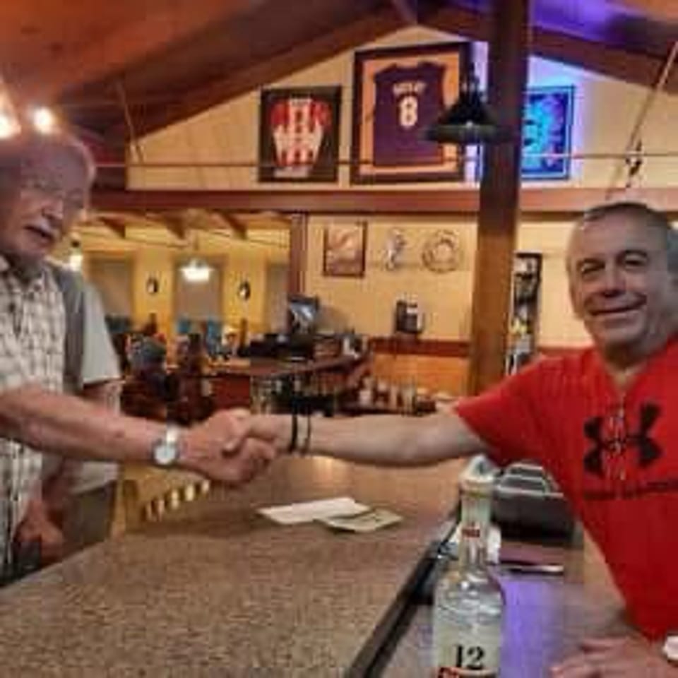 Two men shake hands across a restaurant bar surrounded by warm wood decor, framed sports memorabilia, and drinks.