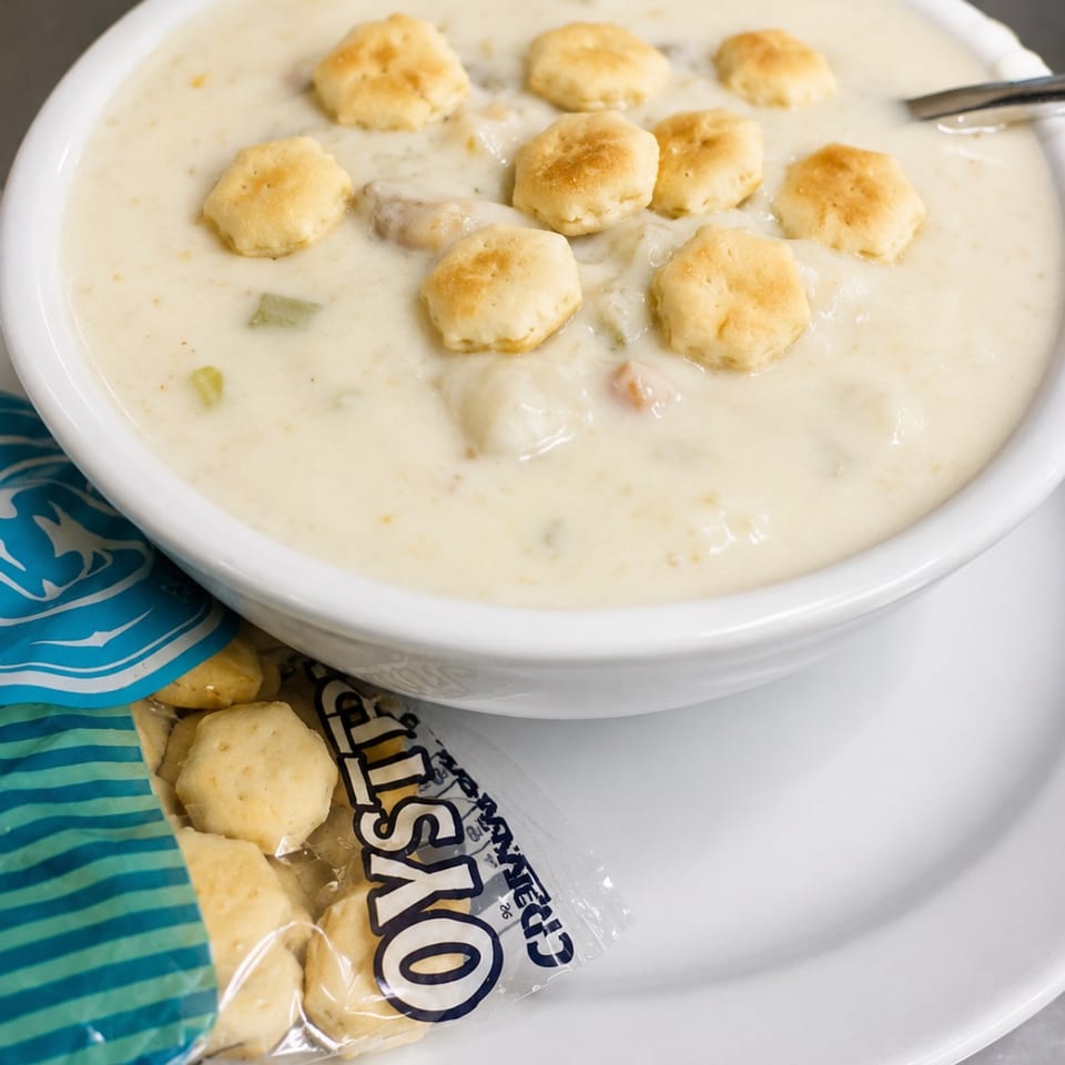 Creamy clam chowder in a white bowl topped with oyster crackers, served with a cracker packet.