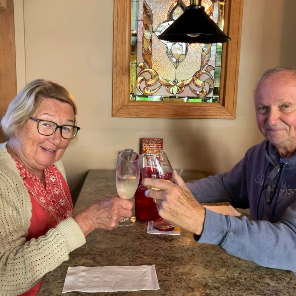 Two diners toast with drinks at a restaurant table beneath a stained-glass window and pendant lamp.