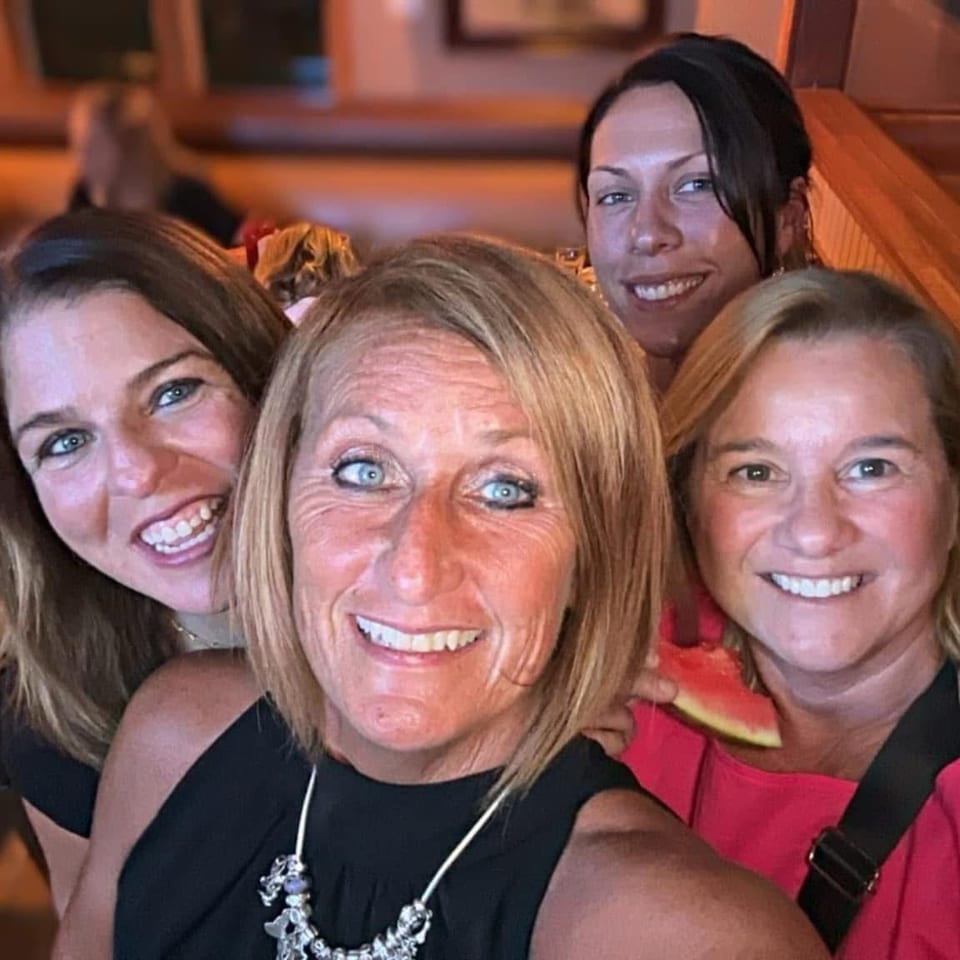 Four smiling women pose together inside a warmly lit restaurant, with wooden booths and other diners visible in the background.