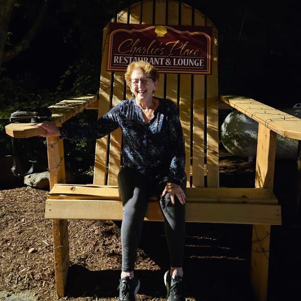 Smiling woman seated on a large wooden chair beneath a Charlie’s Place Restaurant & Lounge sign outdoors at night.