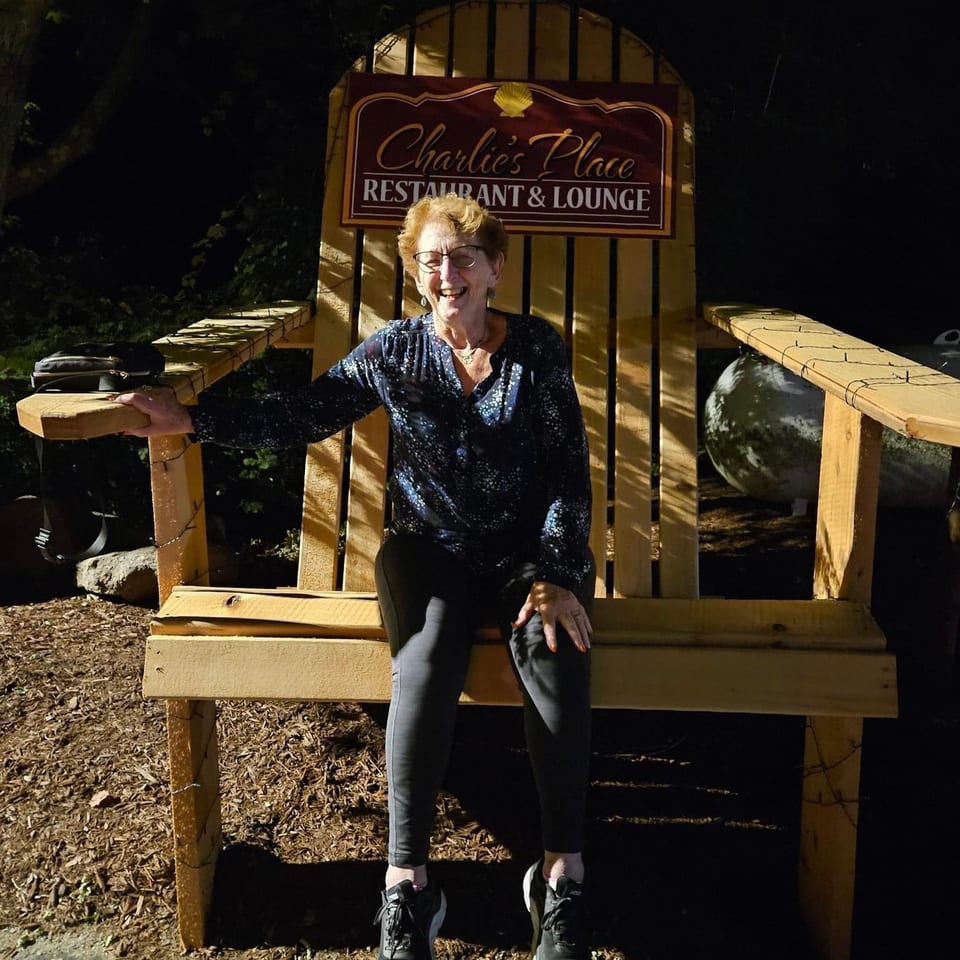 Smiling woman seated on a large wooden chair beneath a Charlie’s Place Restaurant & Lounge sign outdoors at night.