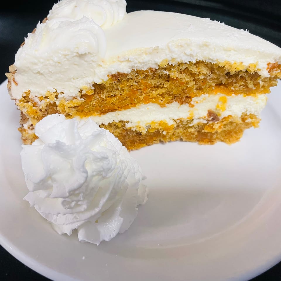 Slice of carrot cake with cream cheese frosting and whipped cream on a white plate.