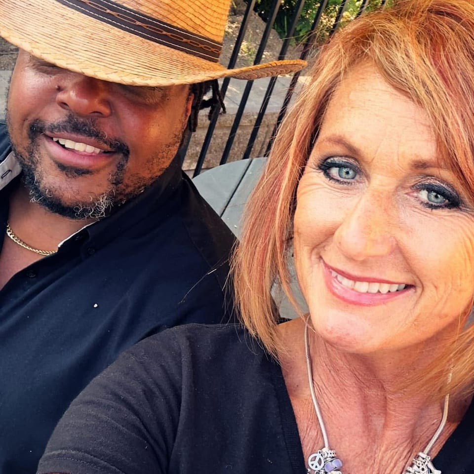 Smiling couple takes a selfie outdoors beside a railing and greenery, wearing black shirts and a wide-brimmed straw hat.