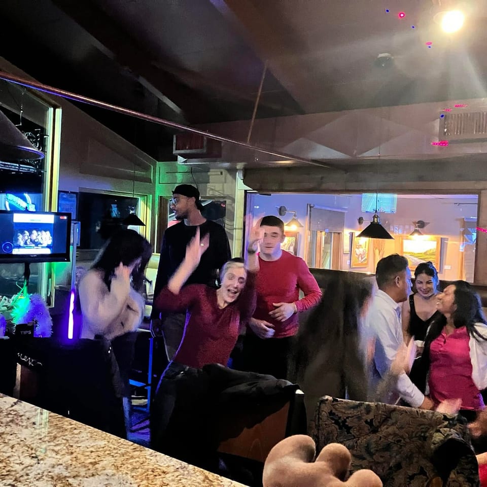 Restaurant patrons gather and socialize beneath colorful lights near a bar and DJ booth.