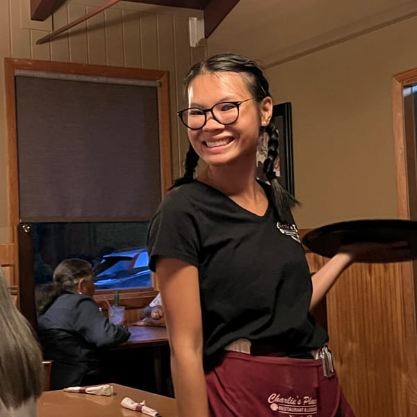 Smiling restaurant server wearing glasses and a burgundy apron stands beside a dining table in a warmly lit dining room.