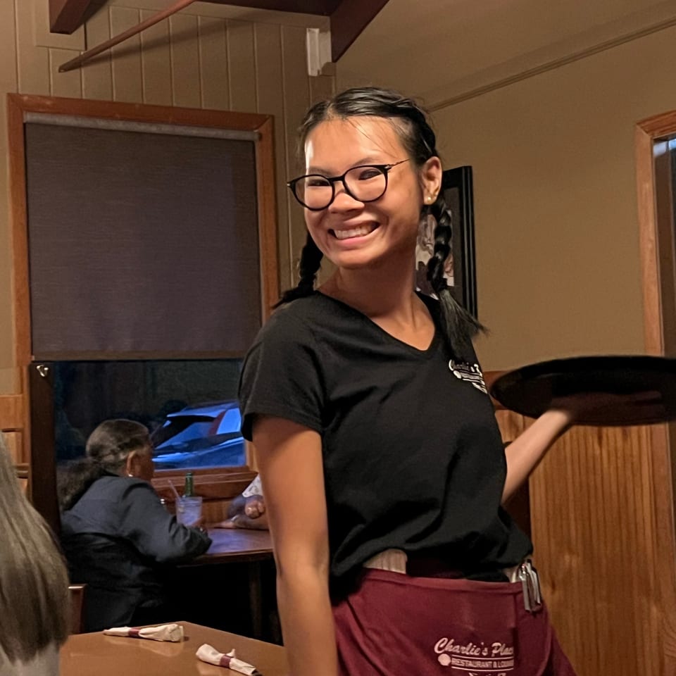 Smiling restaurant server wearing glasses and a burgundy apron stands beside a dining table in a warmly lit dining room.
