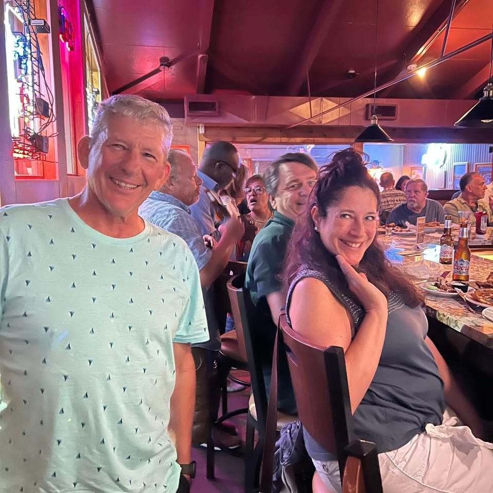 Smiling diners pose inside a lively restaurant, surrounded by tables with meals, drinks, and colorful ambient lighting.