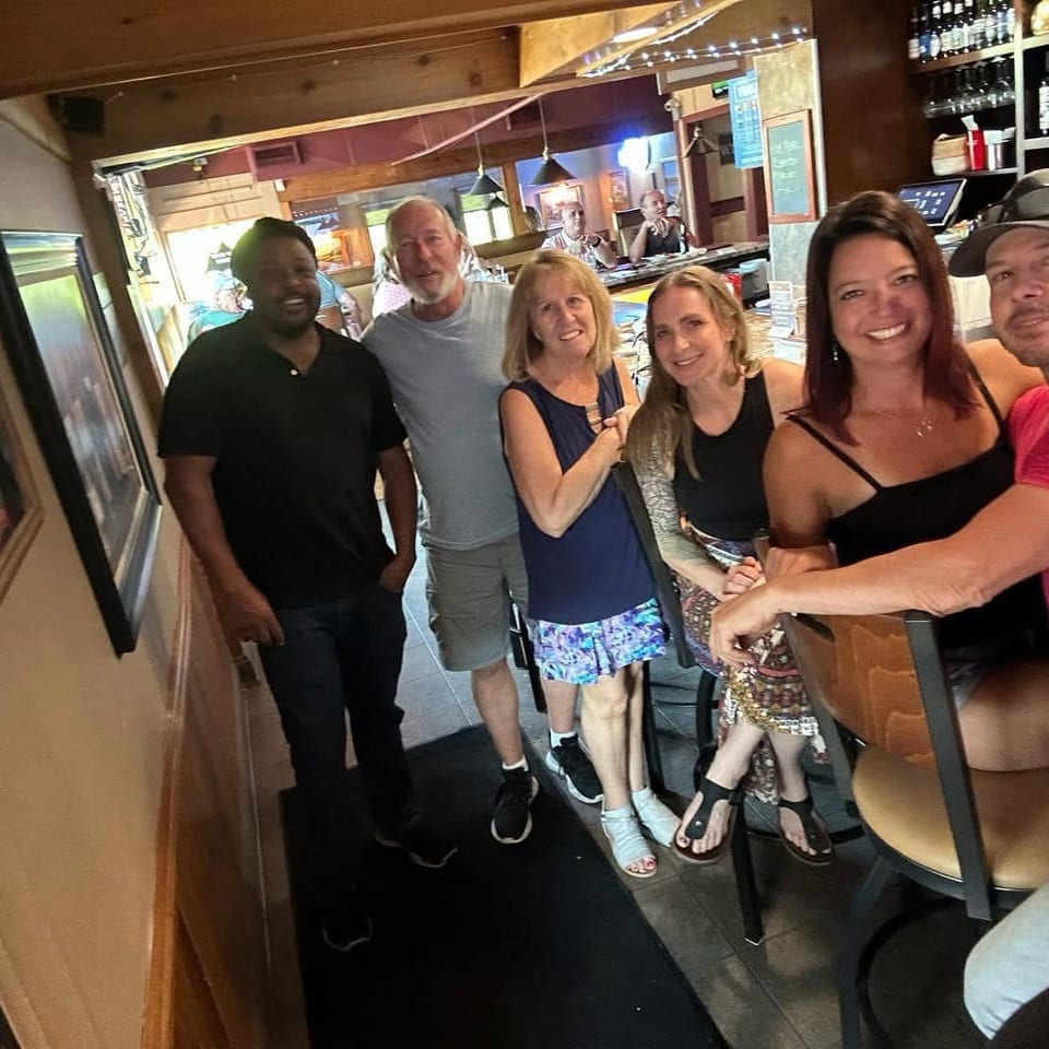 Six smiling adults pose together inside a warmly lit restaurant bar with wood interiors and shelves of bottles.