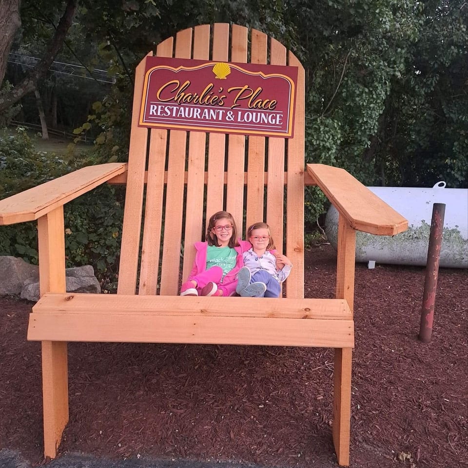 Two children sit on an oversized wooden chair beneath a Charlie’s Place Restaurant & Lounge sign, surrounded by trees.
