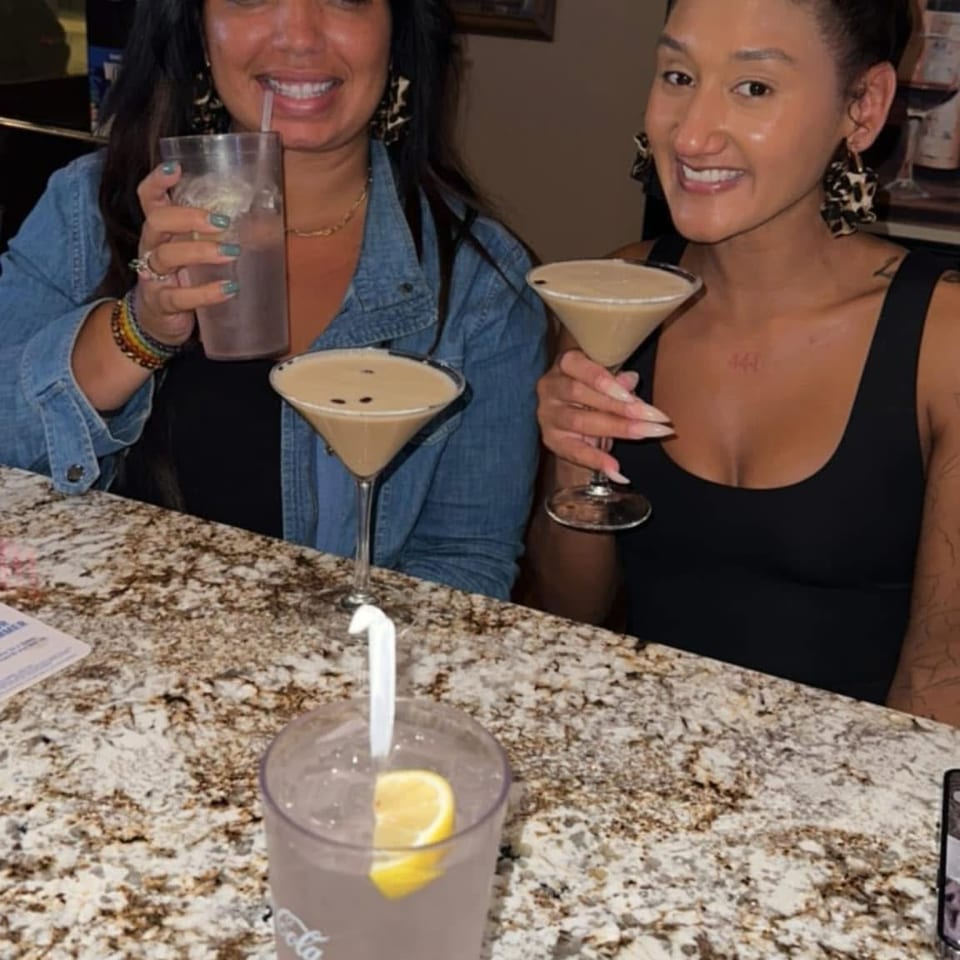Two women smile at a restaurant bar, holding creamy martinis beside a tall drink on a granite countertop.