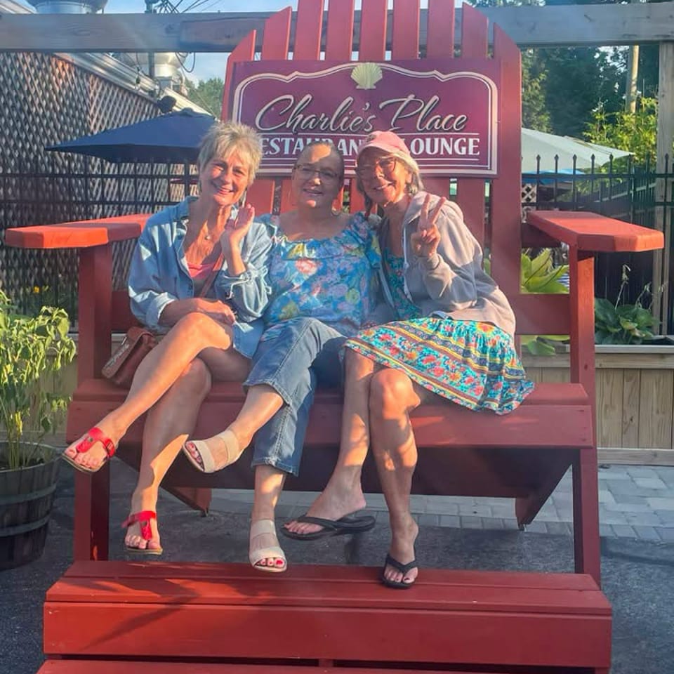 Three women pose on a red bench outside Charlie’s Place Restaurant and Lounge, with patio seating and greenery surrounding them.