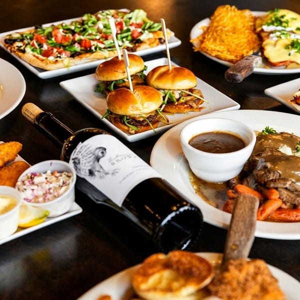 A table laden with various dishes including breakfast, sliders, pizza, and pot roast, with a wine bottle.