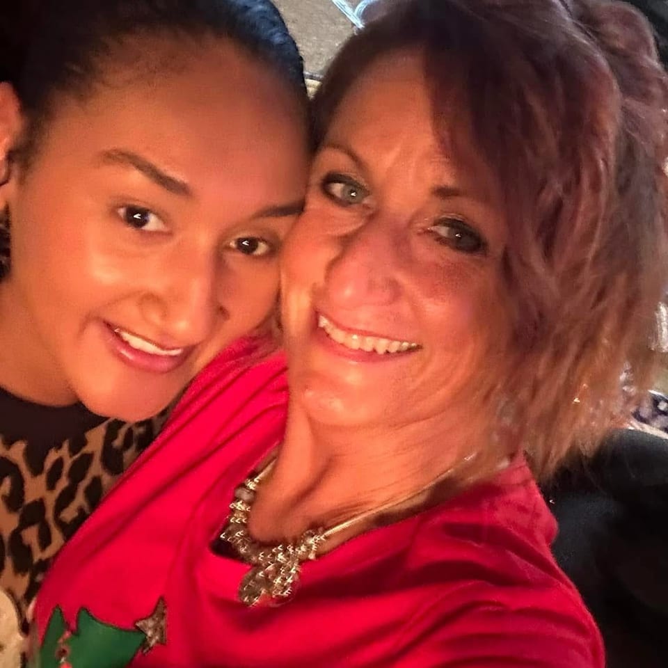 Two smiling women pose closely for a selfie in a warmly lit restaurant, with a plate of food and dining supplies behind them.