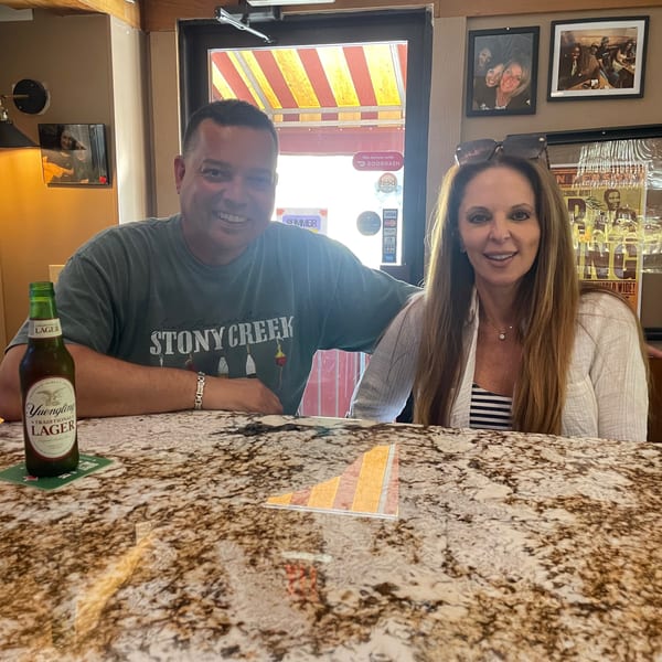 Two smiling diners sit at a restaurant bar with beer and a mixed drink, surrounded by framed photographs and memorabilia.