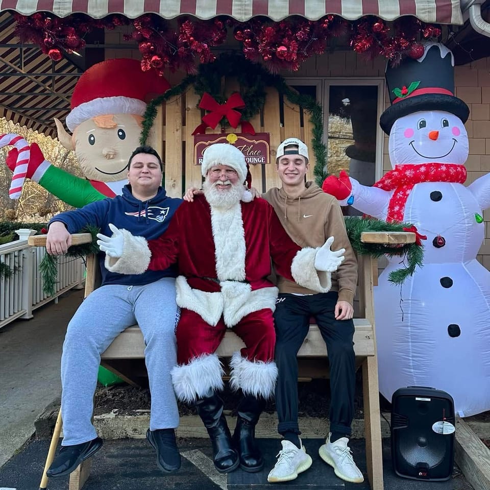 Two people pose with Santa on a bench outside a decorated restaurant, flanked by inflatable Christmas elf and snowman figures.