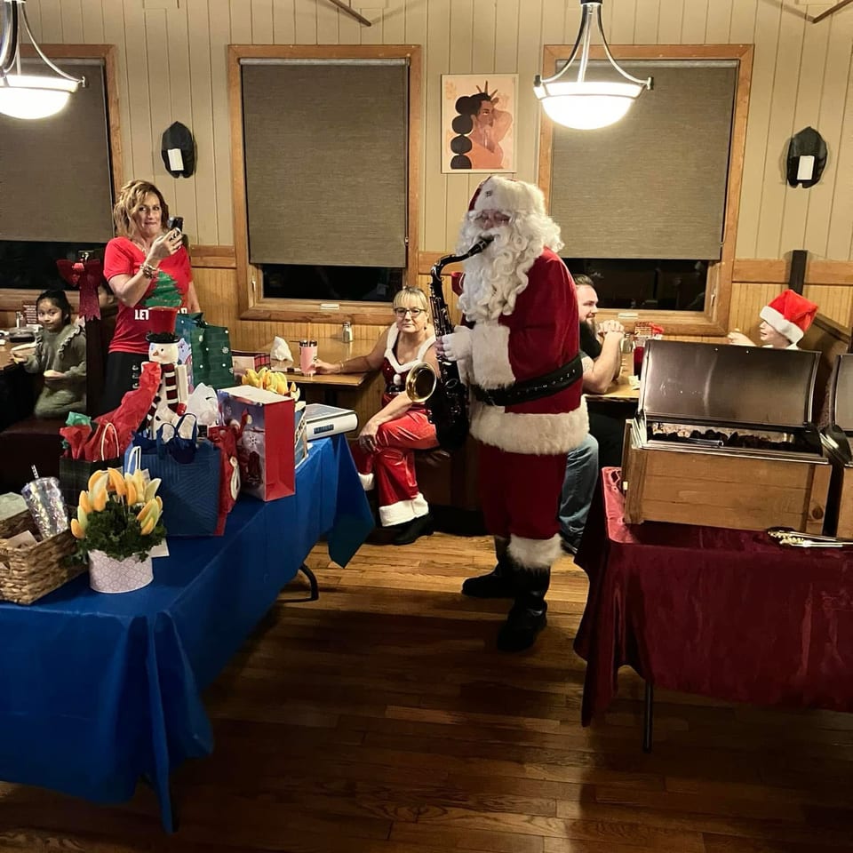 Santa Claus plays a saxophone among diners in a warmly lit restaurant decorated with gifts and holiday decorations.
