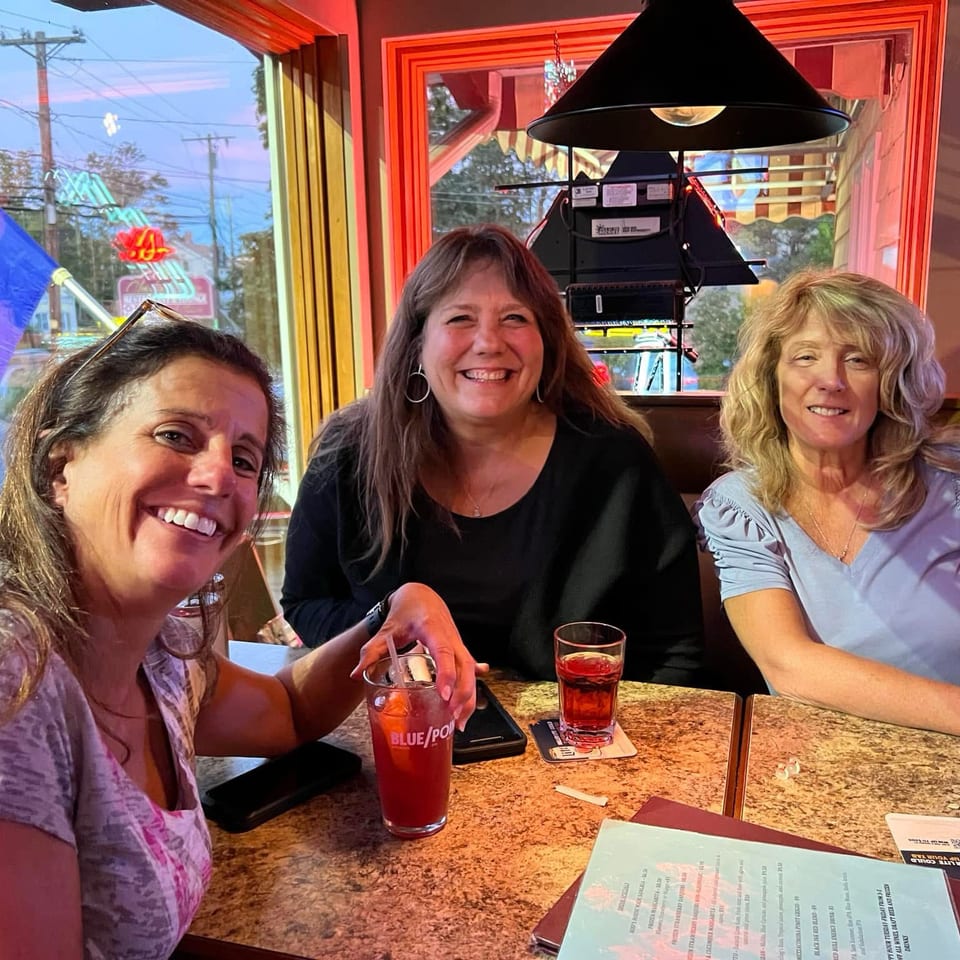 Three smiling women sit together at a restaurant table with drinks and menus, beneath a large hanging lamp.