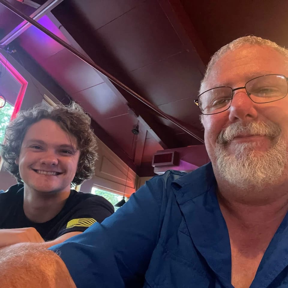 Two smiling people pose for a selfie inside a warmly lit restaurant with colorful neon lighting.