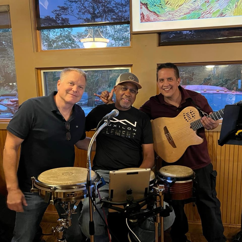 Three musicians pose indoors with drums, a guitar, microphones, and music equipment near restaurant windows.