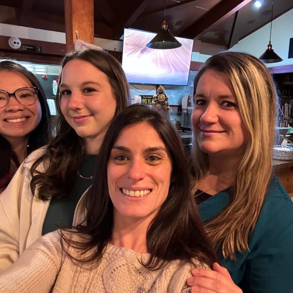 Four smiling women take a selfie inside a warmly lit restaurant near a buffet counter and hanging pendant lights.