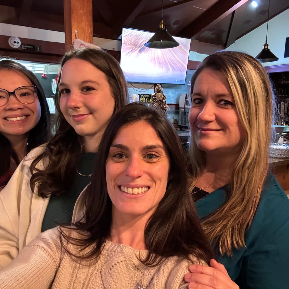 Four smiling women take a selfie inside a warmly lit restaurant near a buffet counter and hanging pendant lights.
