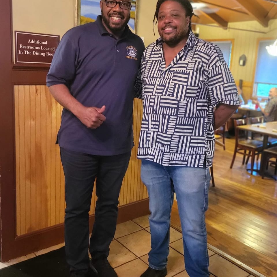 Two smiling men pose inside a warmly lit restaurant near a sign directing guests to additional restrooms.